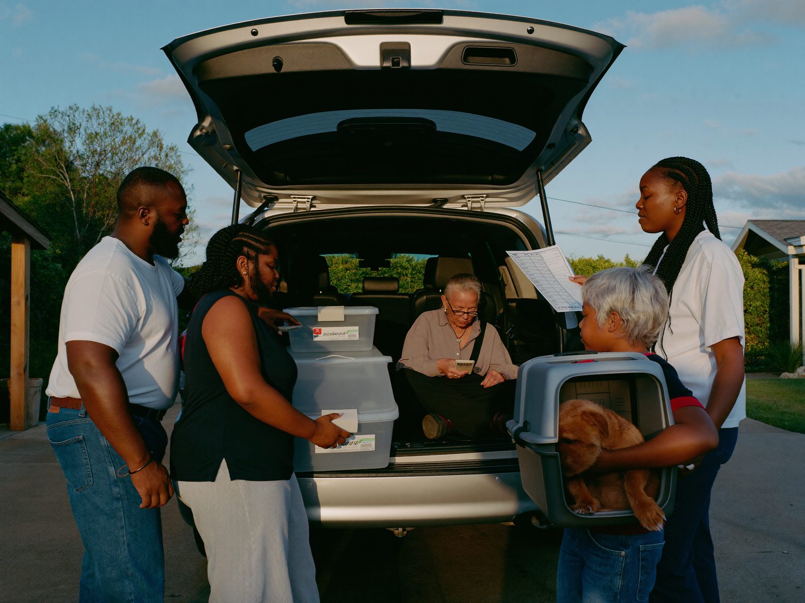 A family calmly loading labeled bins, a checklist, and a pet carrier into their minivan at golden hour